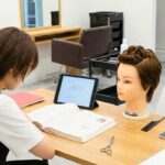 A cosmetology student studying for their state board exam with a textbook and mannequin head in a professional salon training environment.