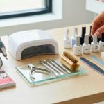 A meticulously organized collection of professional nail technology tools and equipment resting on a clean, light wood desk. The setup includes essential permanent items: a high-torque electric nail file (E-file) machine with various bits, a modern white LED/UV curing lamp, and sterile stainless steel implements like cuticle nippers and pushers resting on a glass tray. Adjacent to these are consumable materials including files, buffers, a wooden-handled nail brush, lint-free wipes, and a notepad. A collection of unbranded professional gel polish, primer, and base coat bottles are lined up on the right, with a manicured hand reaching to select one.