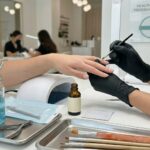 A professional nail technician wearing black gloves and a mask performs a service at a clean workstation with sterilized tools, a UV lamp, and aftercare products.