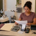 Independent nail salon operator reviewing business folders, receipts, a calculator, a payment terminal, appointment records, keys, and professional nail equipment at a reception counter.