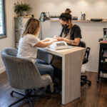 Independent nail technician serving a client in a compact salon booth with a manicure table, rolling storage, task lighting, organized supplies, and dust ventilation.