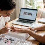 New nail technician practicing a manicure at a home desk with a laptop, organized nail kit, and job application materials nearby.