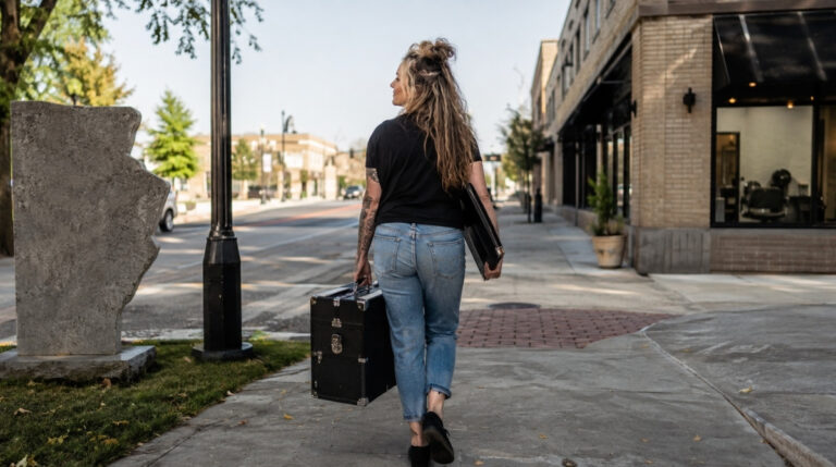 A cosmetologist walks toward a salon in a new downtown neighborhood while carrying a hairstyling case and professional document portfolio.