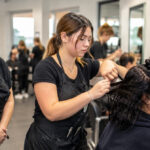 Cosmetology student works on a real client while an experienced instructor observes the hair sectioning and styling process in a busy training salon.