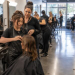 Cosmetology student works on a client’s hair while an instructor supervises in an active training salon with mannequin and beauty practice stations nearby.