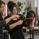 Seasoned salon professional points to a mannequin hair section as a student holds a comb and shears while a supervising instructor observes.
